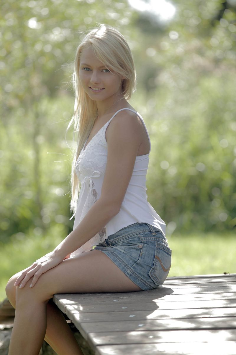 cute young blonde girl with blue eyes reveals her white camisole and jean skirt outside on the small wooden bridge