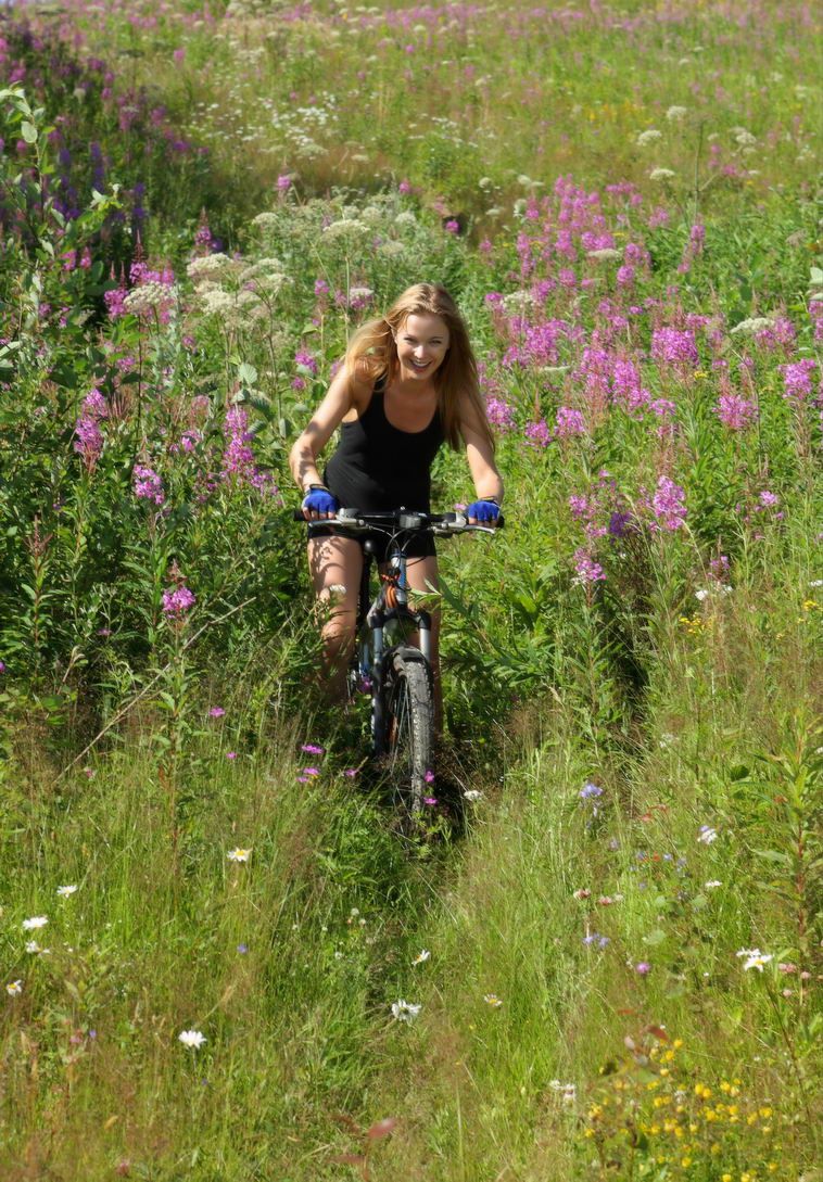 cute young blonde girl reveals on the field of wild flowers with a bicycle