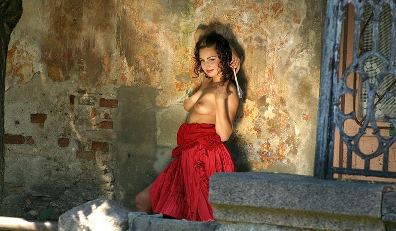 young curly brunette girl reveals in a red dress at the stone building entrance