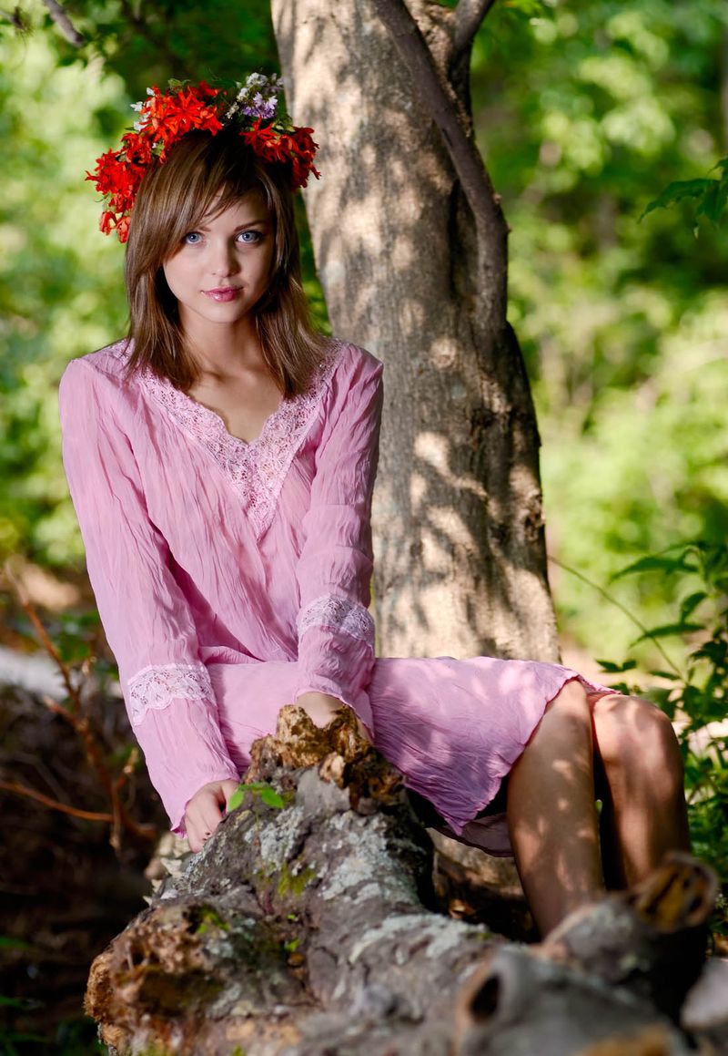 young dark blonde girl with blue eyes and a floral wreath reveals a pink nightgown in the forest