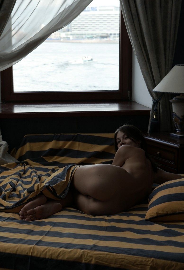 young brunette girl near the hotel room window posing on the bed with striped sheets