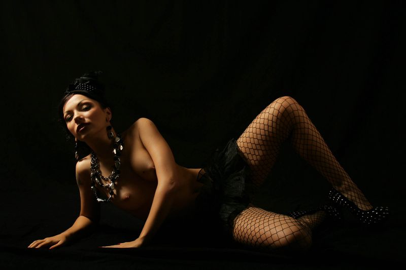 black haired girl with a necklace and earrings undresses her black coat, skirt and fishnet stockings in the studio
