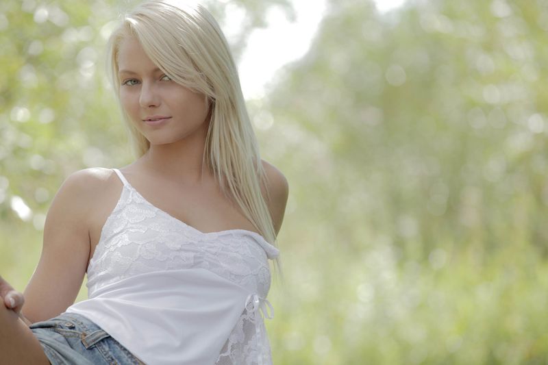 cute young blonde girl with blue eyes reveals her white camisole and jean skirt outside on the small wooden bridge