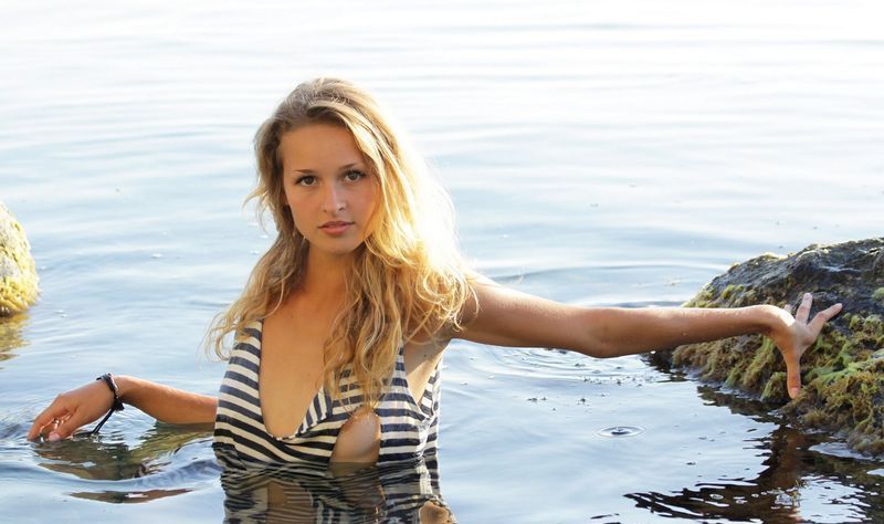 young blonde girl with tan lines reveals her black and white striped sleeveless tank top shirt on the rocky shore at the sea